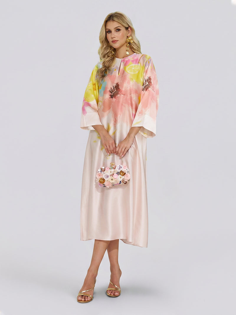 A woman wearing the Social Queen Aquarelle Blooms 100% Mulberry Silk Dress, paired with a matching floral handbag and open-toe heels, stands against a plain light background—an elegant choice for a wedding guest ensemble.