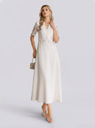 A woman models the Social Queen Lace-Panel Satin A-Line Gown—an elegant ivory dress featuring a graceful A-line silhouette and a delicate lace-paneled bodice. She accessorizes with a small handbag and stands against a plain light background.