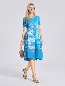 A woman wears the Social Queen Lake Blue Print Plissé A-Line Dress—featuring short sleeves and a knee-length, watercolor-inspired floral print—paired with gold heels and a small handbag, standing against a plain light background.