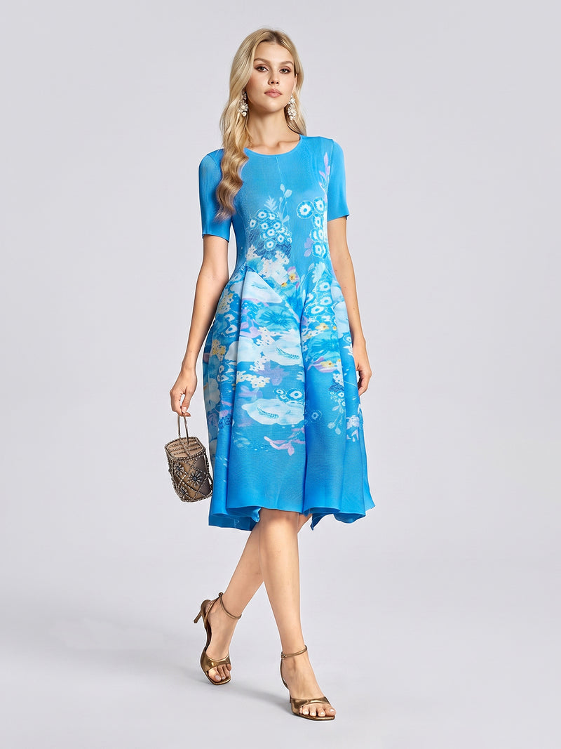 A woman wears the Social Queen Lake Blue Print Plissé A-Line Dress—featuring short sleeves and a knee-length, watercolor-inspired floral print—paired with gold heels and a small handbag, standing against a plain light background.