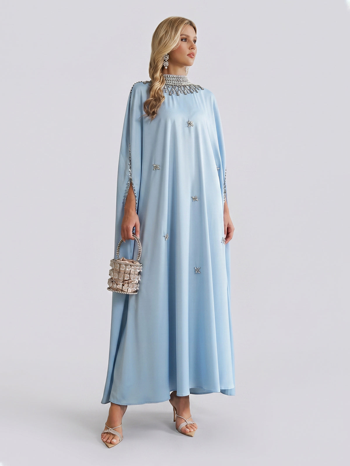 A woman stands against a plain light background, wearing the Social Queen Crystal-Embellished Mock-Neck Caftan Dress in light blue, paired with heels and a small silver handbag.