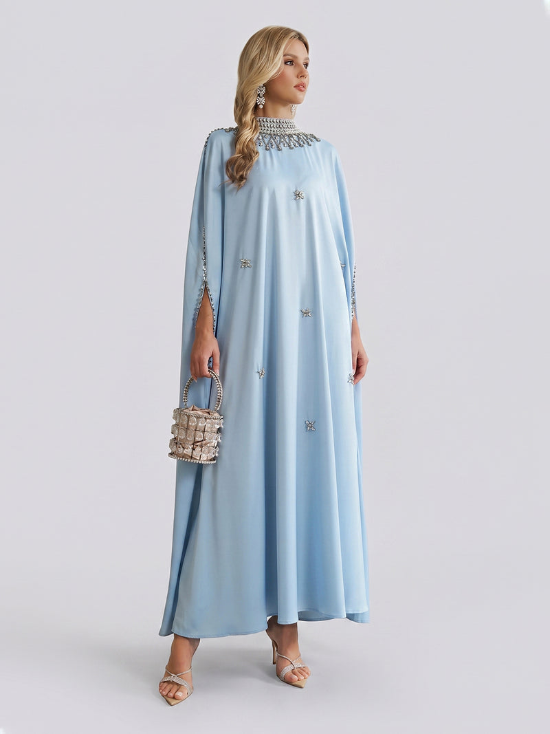 A woman stands against a plain light background, wearing the Social Queen Crystal-Embellished Mock-Neck Caftan Dress in light blue, paired with heels and a small silver handbag.