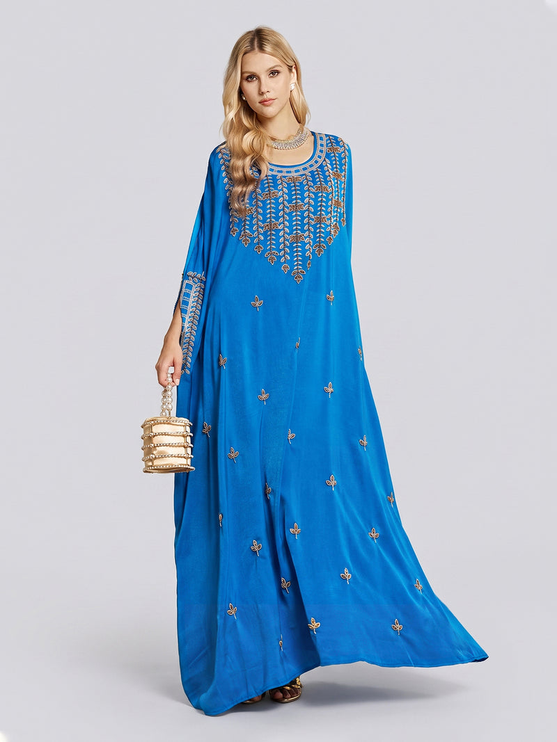 A woman stands against a plain light background, wearing the Social Queen Gilded Leaf Embellished Cape Dress in bright royal blue chiffon satin with intricate gold leaf embroidery. She holds a gold bag, wears a silver necklace, and her long blonde hair is styled in loose waves.