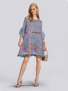 A woman wearing the Social Queen Art-Print Floral-Appliqué Pleated Dress with ruffled sleeves, paired with metallic heels and carrying a small woven bag, stands against a plain background.