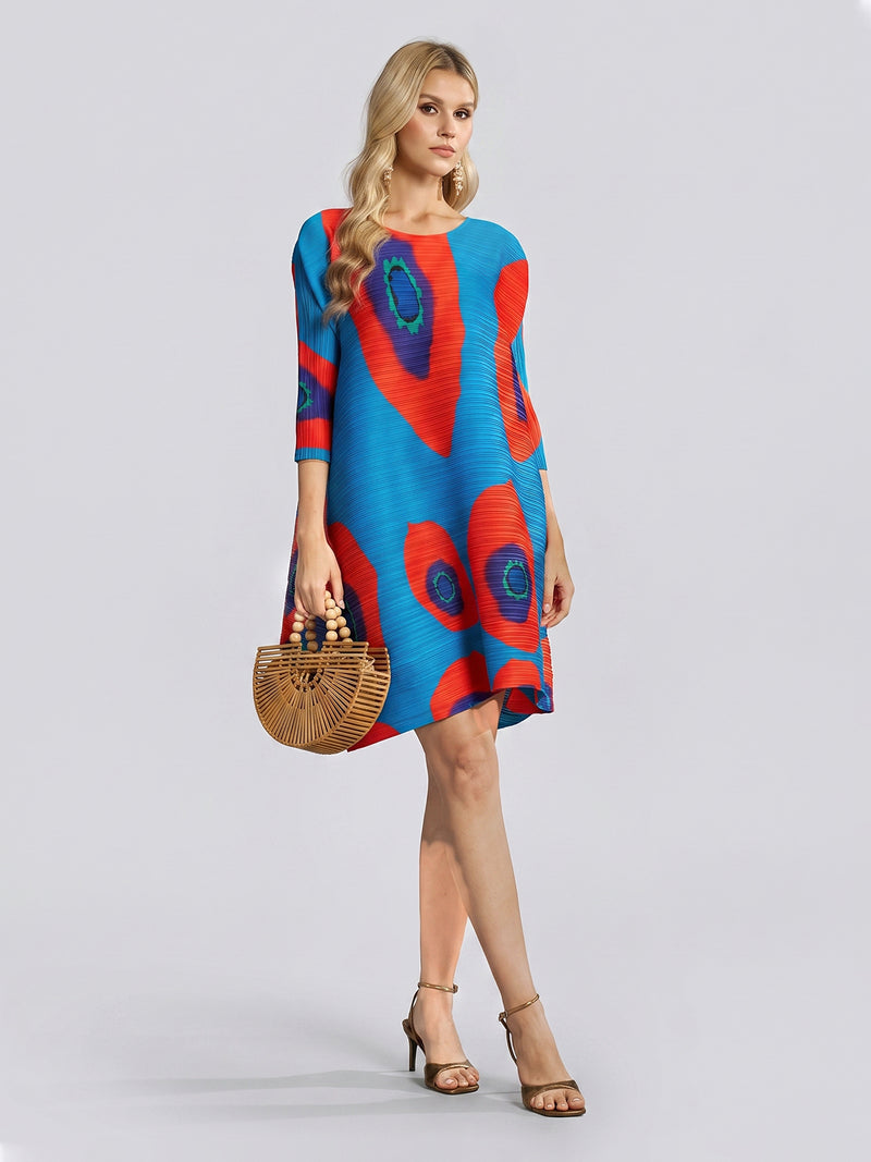 A woman in the Social Queen Artistic Print Plissé A-Line Dress, featuring blue fabric with red and green abstract designs, holds a wooden handbag and wears sandals while standing against a plain gray background.