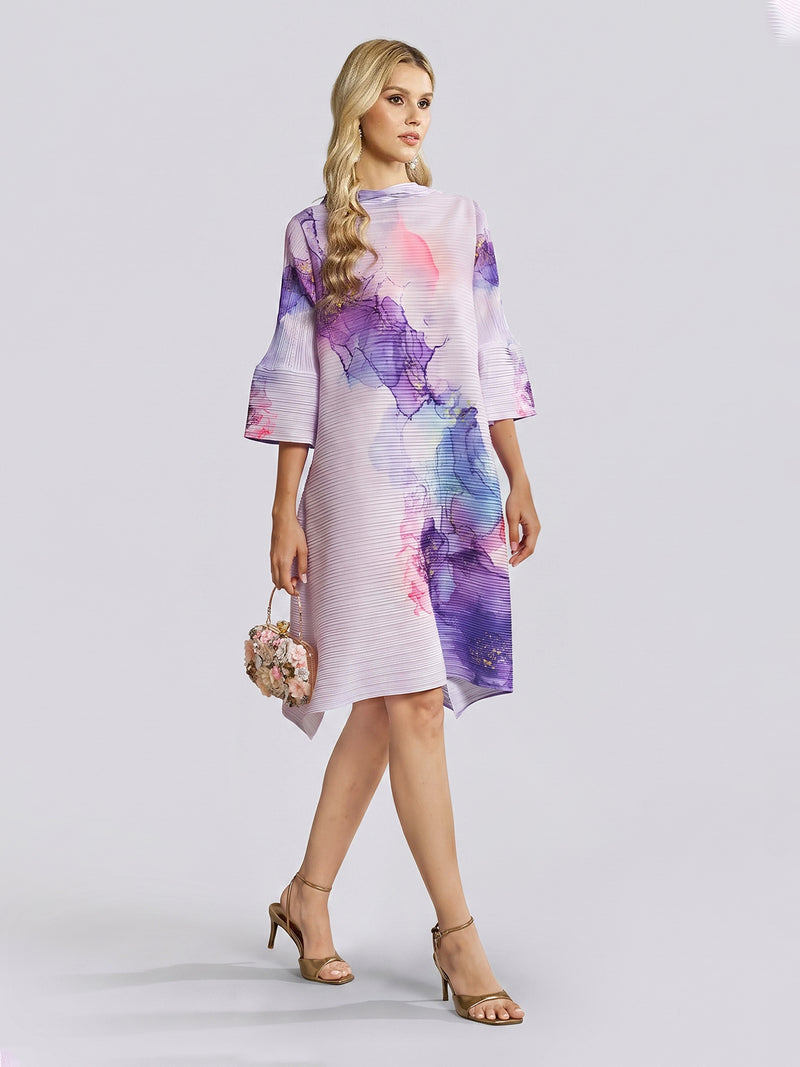 A woman wearing the Social Queen Watercolor Print Plissé Dress, featuring pastel purple and pink florals with bohemian bell sleeves, stands against a plain light background. She holds a round floral purse and wears beige sandals.