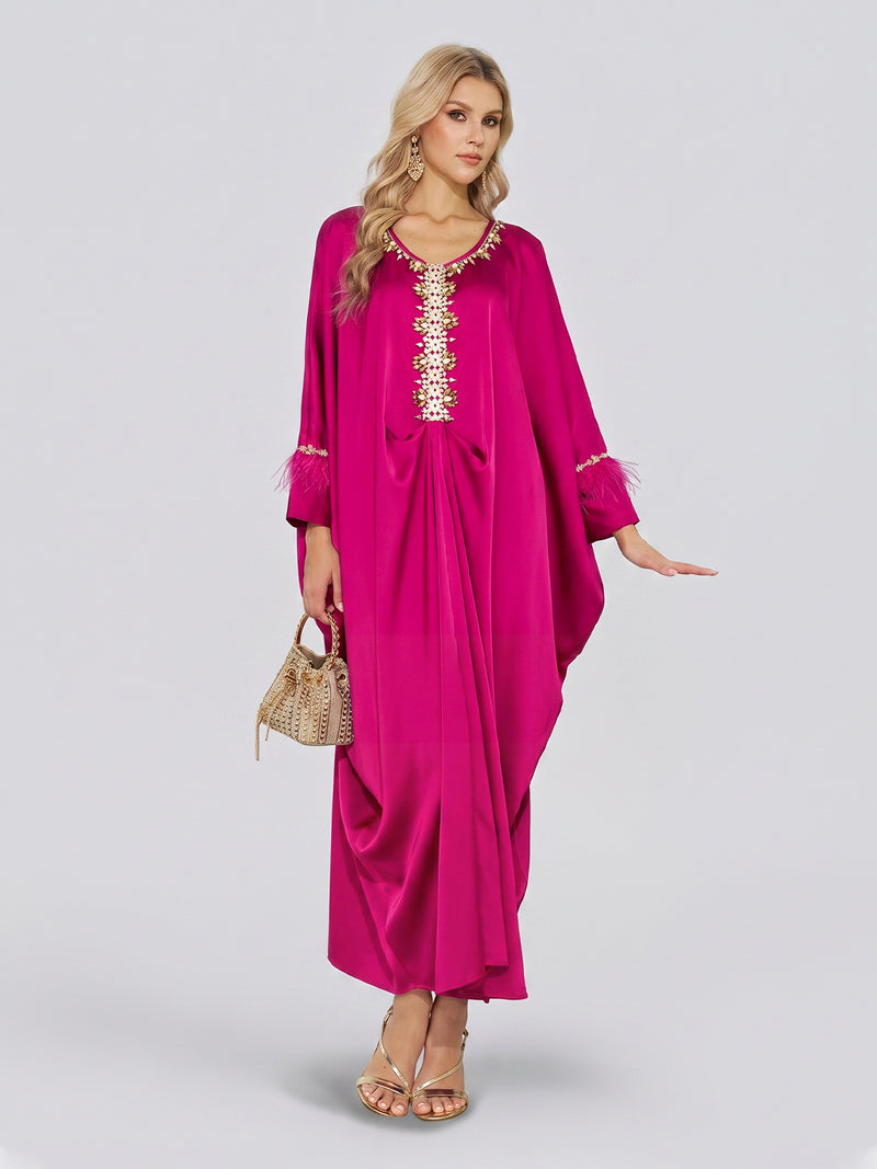 A woman poses against a plain light background wearing the Social Queen Feather-Trimmed Embellished Satin Caftan Dress— a long, fuchsia satin gown adorned with a beaded neckline, gold embroidery, and feathered cuffs—styled with gold sandals and a woven handbag.