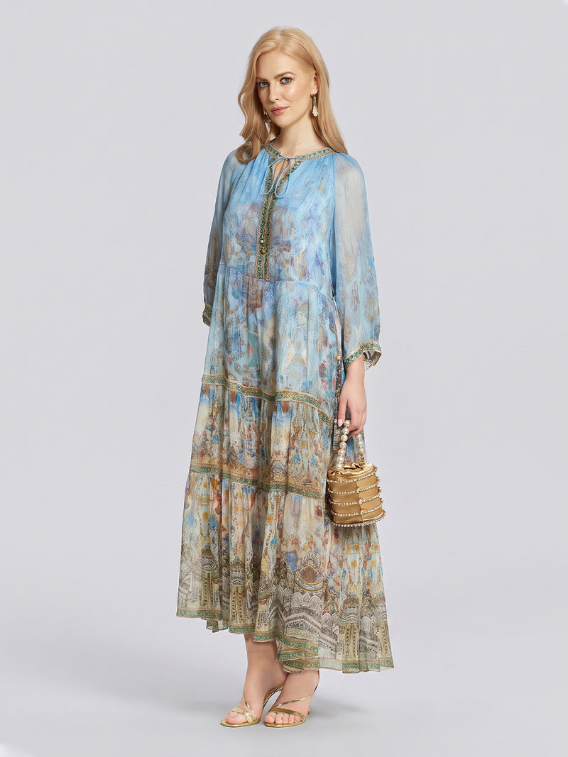 A woman wearing the Social Queen Vintage Palatial Print Tiered Silk Dress, paired with gold earrings, sandals, and a round beaded handbag, stands against a plain light gray background.