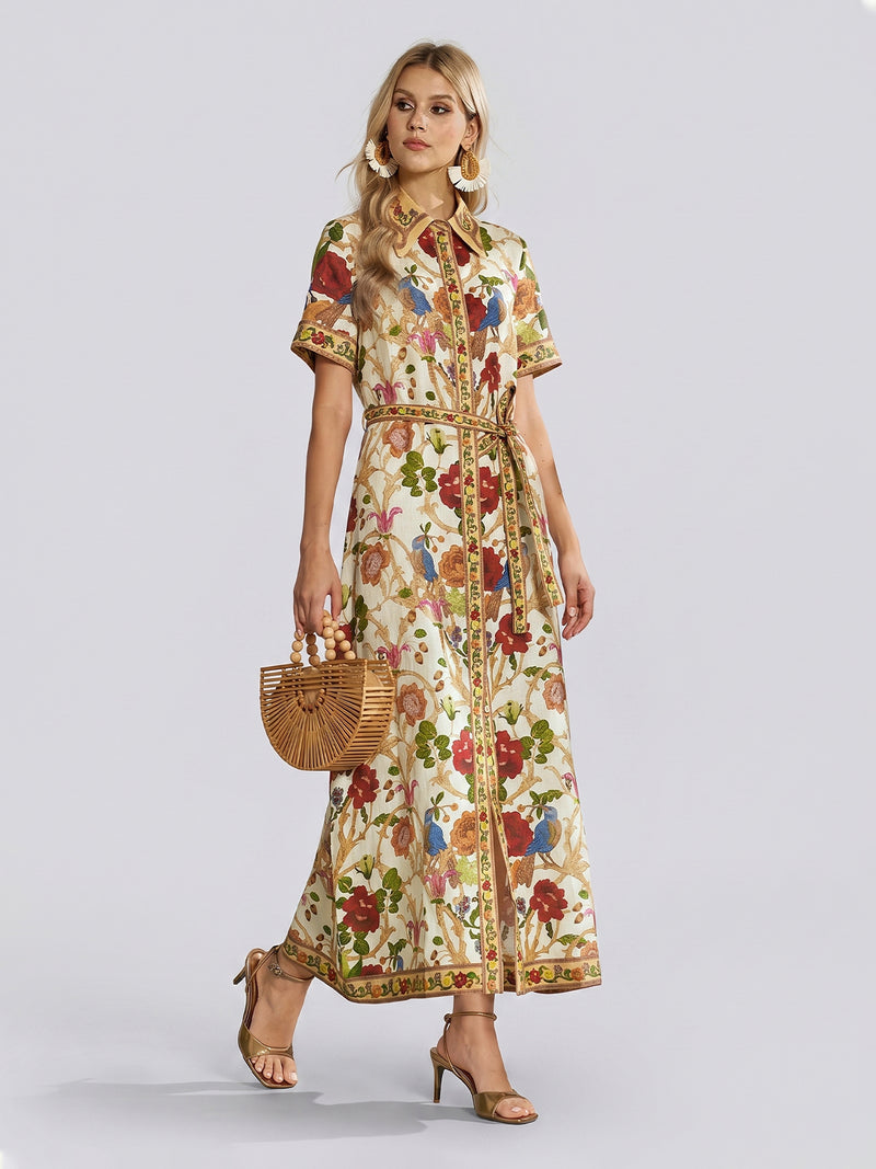 A woman wears the Social Queen Vintage Floral & Bird Print Shirt Dress—featuring short sleeves and a belted waist—with gold sandals and a wooden handbag, standing against a plain light background for an effortlessly chic resort wear look.
