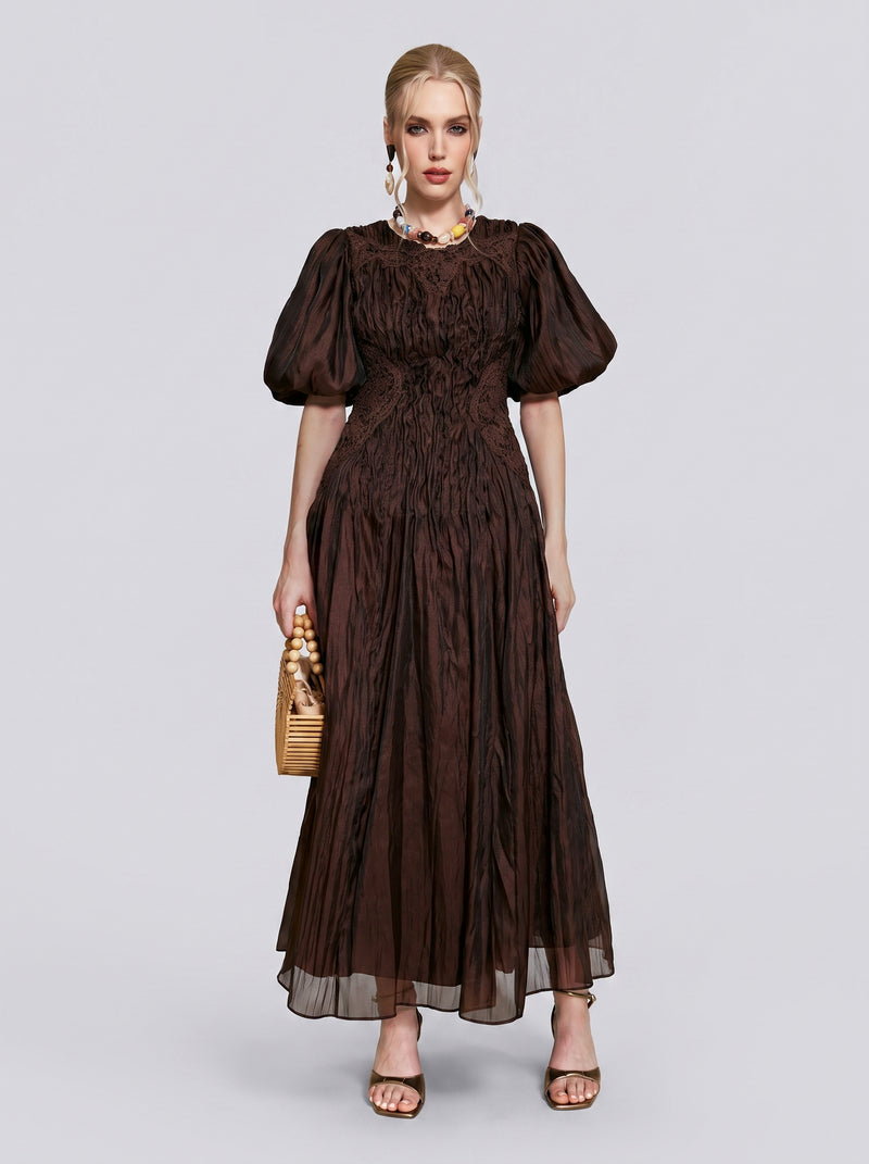 A woman stands against a plain light background wearing the Social Queen Puff Sleeve Textured Lace Dress, which features brown hues, puff sleeves, a gathered bodice, and a fitted A-line silhouette. She accessorizes with statement earrings, sandals, and carries a wicker handbag.