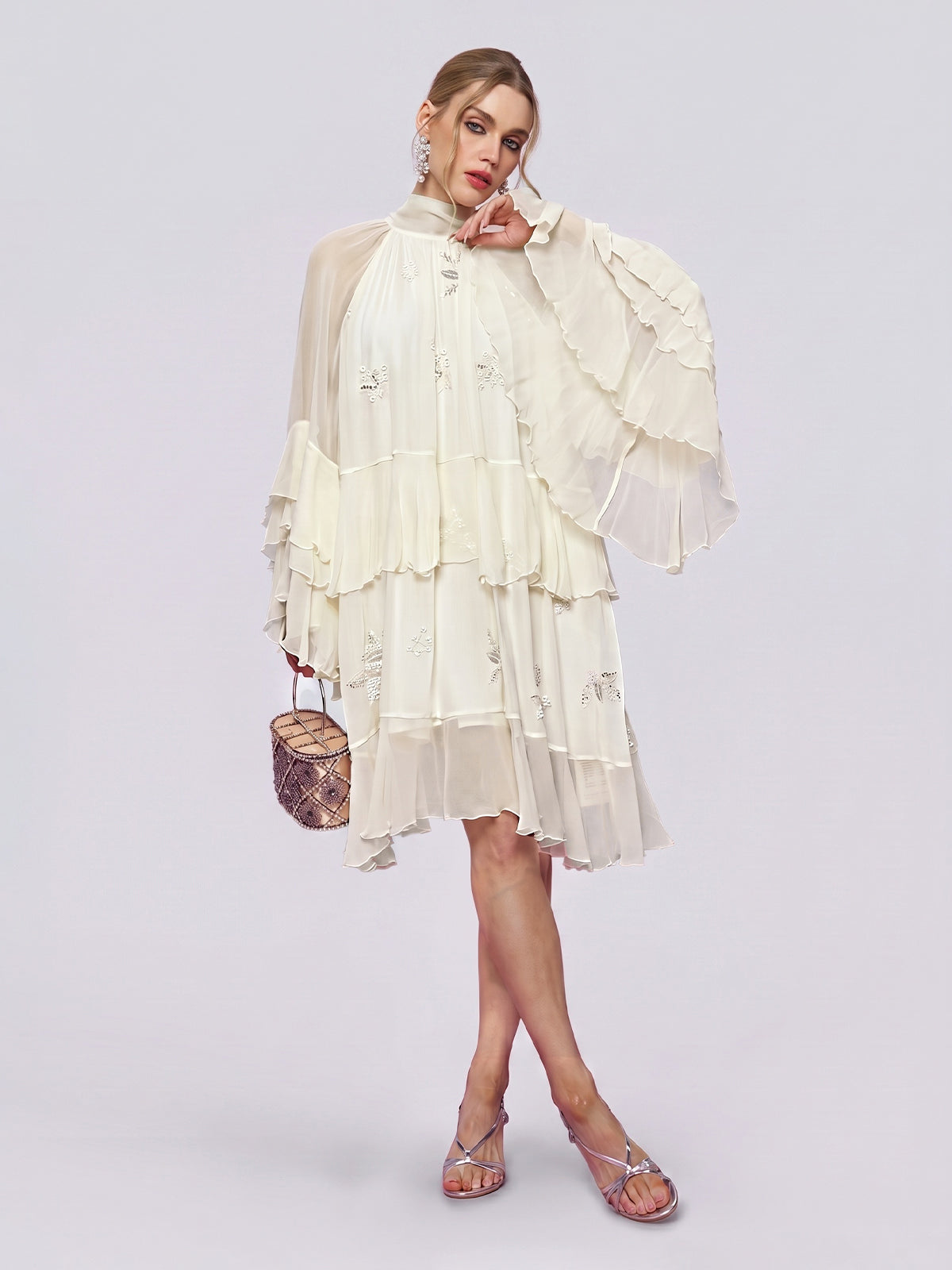 A woman stands against a plain light background wearing the Social Queen Embroidered High-Neck Silk Dress—a tiered, long-sleeve white mulberry silk dress with ruffled details—paired with silver sandals and holding a small brown handbag.