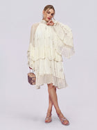 A woman stands against a plain light background wearing the Social Queen Embroidered High-Neck Silk Dress—a tiered, long-sleeve white mulberry silk dress with ruffled details—paired with silver sandals and holding a small brown handbag.