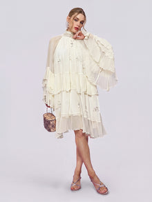 A woman stands against a plain light background wearing the Social Queen Embroidered High-Neck Silk Dress—a tiered, long-sleeve white mulberry silk dress with ruffled details—paired with silver sandals and holding a small brown handbag.