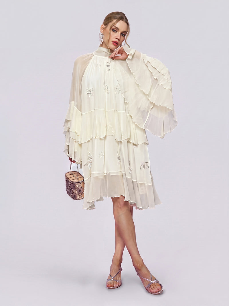 A woman stands against a plain light background wearing the Social Queen Embroidered High-Neck Silk Dress—a tiered, long-sleeve white mulberry silk dress with ruffled details—paired with silver sandals and holding a small brown handbag.