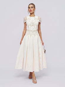 A woman wearing the Appliqué Pearl Waffle Mesh Dress by Social Queen—an ankle-length, white, short-sleeved A-line dress featuring a pleated skirt and intricate pearl embellishments on the bodice—holds a small purse as she stands against a plain light background.