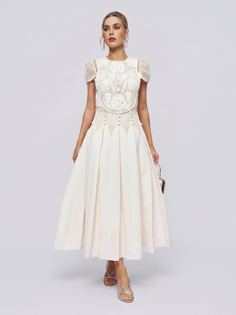 A woman wearing the Appliqué Pearl Waffle Mesh Dress by Social Queen—an ankle-length, white, short-sleeved A-line dress featuring a pleated skirt and intricate pearl embellishments on the bodice—holds a small purse as she stands against a plain light background.