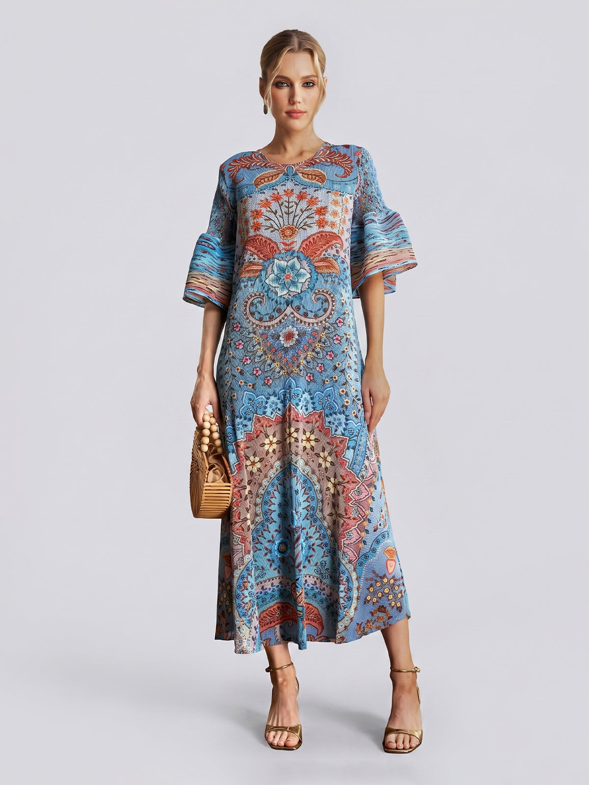 A woman wearing the Social Queen Paisley Print Pleated Midi Dress—featuring flared sleeves, a relaxed silhouette, and a striking blue paisley design—holds a woven handbag and wears heeled sandals while standing against a plain light grey background.