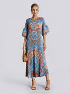 A woman wearing the Social Queen Paisley Print Pleated Midi Dress—featuring flared sleeves, a relaxed silhouette, and a striking blue paisley design—holds a woven handbag and wears heeled sandals while standing against a plain light grey background.