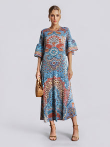 A woman wearing the Social Queen Paisley Print Pleated Midi Dress—featuring flared sleeves, a relaxed silhouette, and a striking blue paisley design—holds a woven handbag and wears heeled sandals while standing against a plain light grey background.