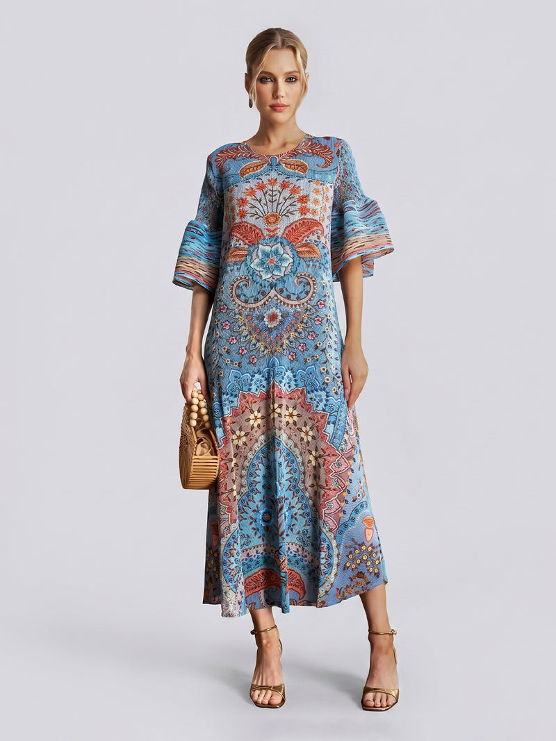 A woman wearing the Social Queen Paisley Print Pleated Midi Dress—featuring flared sleeves, a relaxed silhouette, and a striking blue paisley design—holds a woven handbag and wears heeled sandals while standing against a plain light grey background.