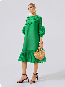A woman stands against a plain background wearing the Social Queen Beaded Floral Pleated Midi Dress in green, paired with high-heeled sandals and holding a wooden handbag.