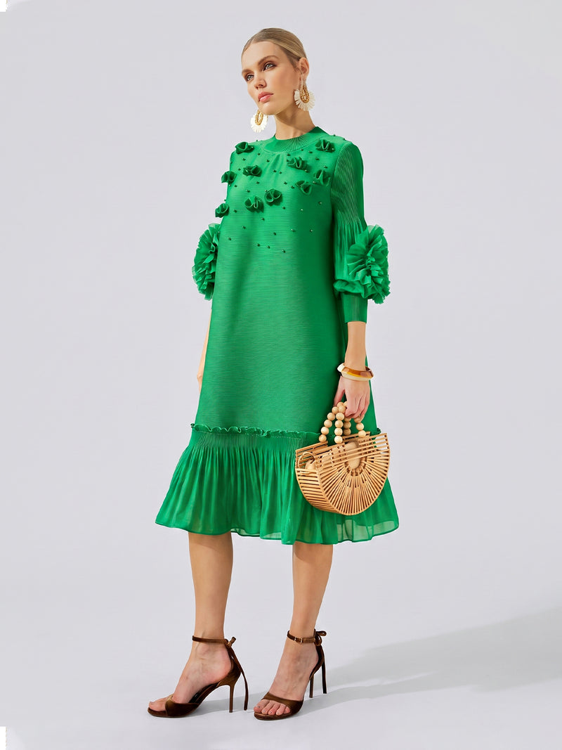 A woman stands against a plain background wearing the Social Queen Beaded Floral Pleated Midi Dress in green, paired with high-heeled sandals and holding a wooden handbag.