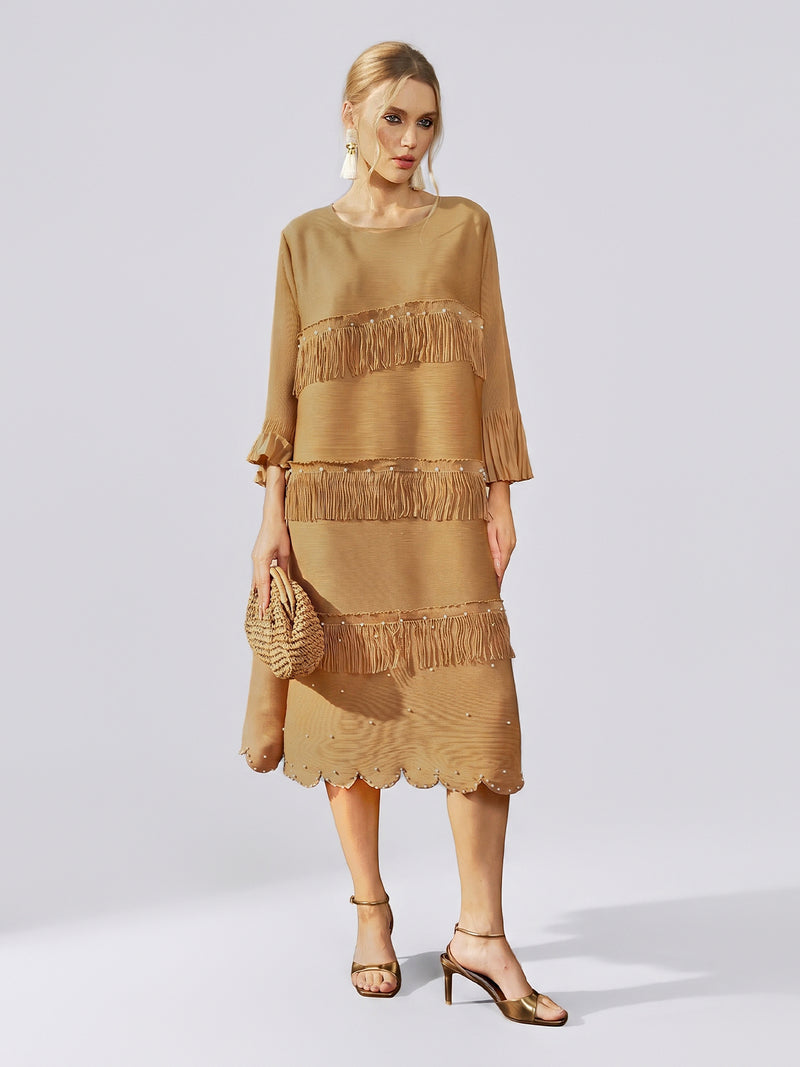 A woman is wearing the Social Queen Pearl Detail Fringe Pleated Dress— a loose, gold, knee-length chiffon dress featuring pleats, layered fringe, pearl details, and a scalloped hem. She pairs it with a woven clutch and gold heels against a plain background.