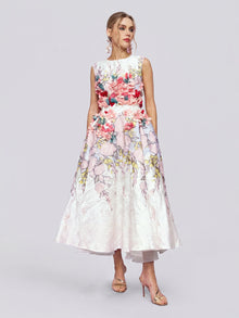 A woman wearing the Social Queen 3D Floral Embellished Jacquard Gown, a sleeveless, mid-length white dress adorned with colorful 3D floral appliqué, stands in heels against a plain light background.