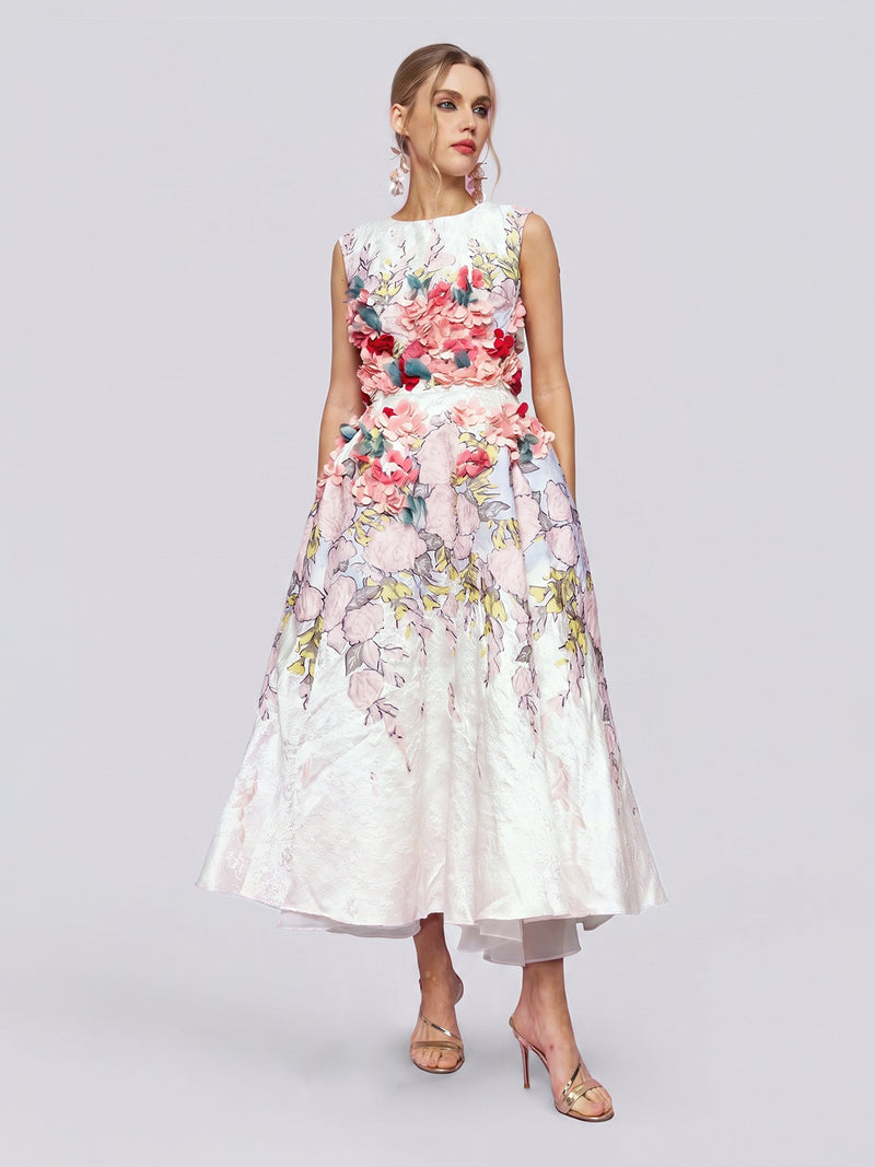 A woman wearing the Social Queen 3D Floral Embellished Jacquard Gown, a sleeveless, mid-length white dress adorned with colorful 3D floral appliqué, stands in heels against a plain light background.