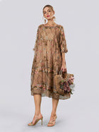 A woman models the Social Queen Square-Neck Embroidered Silk Dress, a sheer, knee-length piece featuring short sleeves and delicate floral embroidery. She holds a woven handbag adorned with flowers and stands in high-heeled sandals against a plain light backdrop.