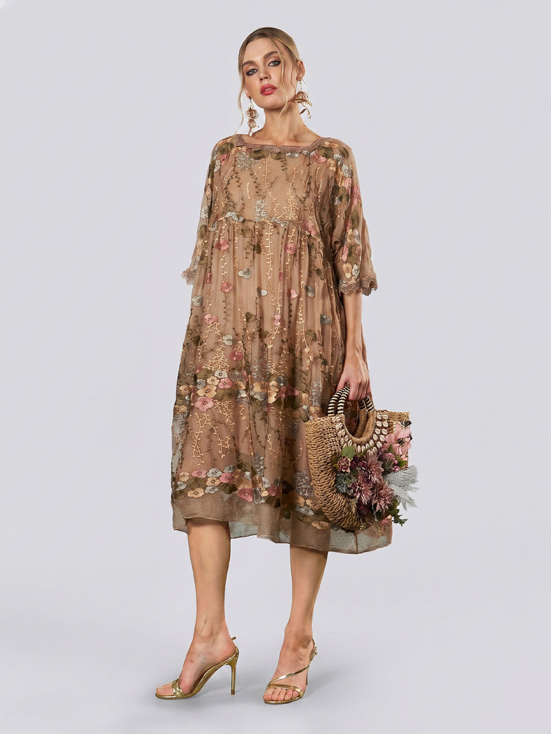 A woman models the Social Queen Square-Neck Embroidered Silk Dress, a sheer, knee-length piece featuring short sleeves and delicate floral embroidery. She holds a woven handbag adorned with flowers and stands in high-heeled sandals against a plain light backdrop.