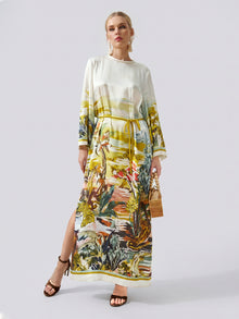 A woman wearing the Social Queen Landscape Print Satin Silk Dress, featuring a vibrant tropical landscape print, holds a wooden bag and stands against a plain light background.