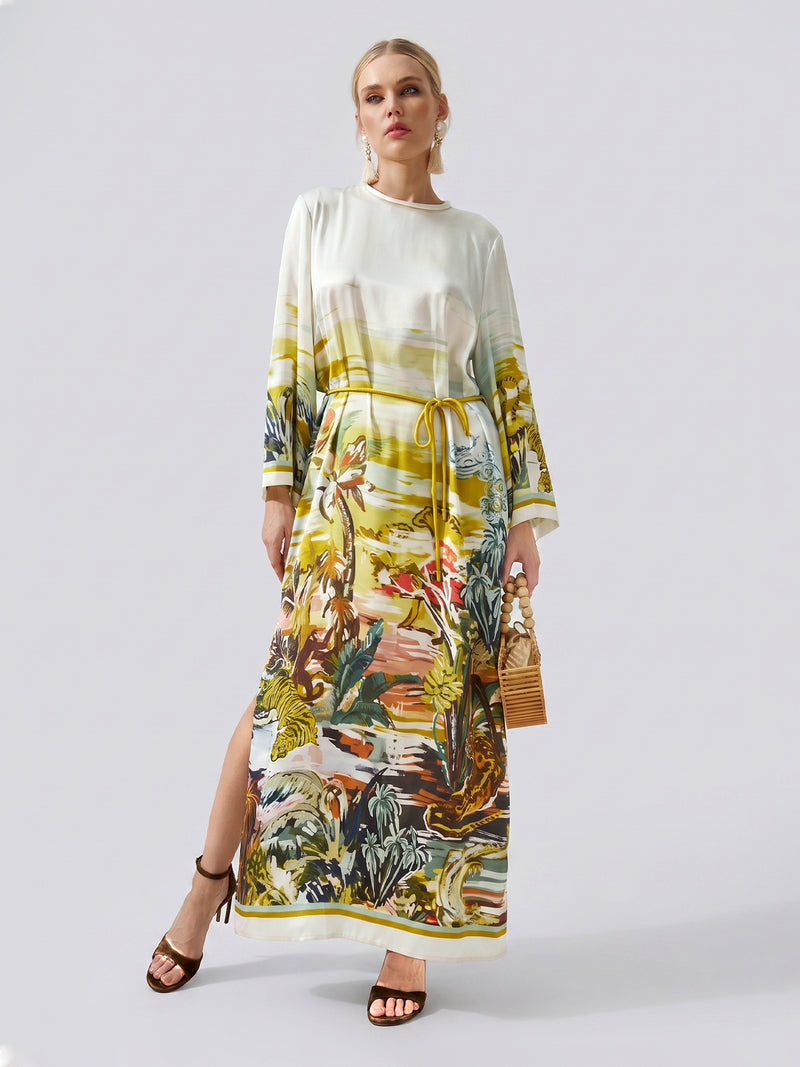 A woman wearing the Social Queen Landscape Print Satin Silk Dress, featuring a vibrant tropical landscape print, holds a wooden bag and stands against a plain light background.