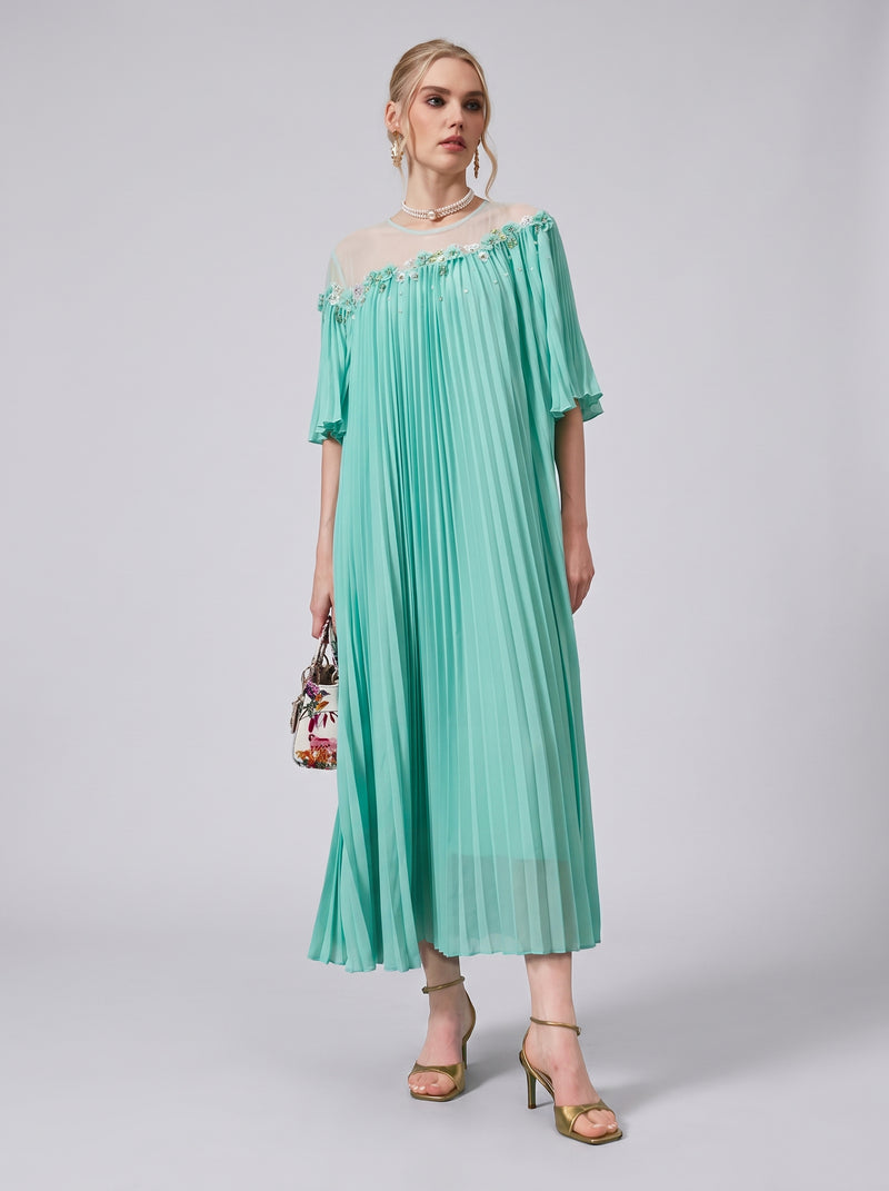 A woman wears the Social Queen Floral Embellishment Pleated Dress in mint green, showcasing a relaxed silhouette with an off-the-shoulder sheer neckline. She carries a floral handbag adorned with delicate beadwork and stands in heels against a plain background.