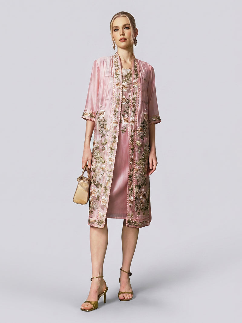 A woman models the Rose Embroidered Tencel Dress Set from Social Queen, featuring a satin-finish slip dress and matching jacket adorned with intricate floral embroidery in soft pink. She pairs the look with gold heels and a beige handbag, standing against a plain light background.