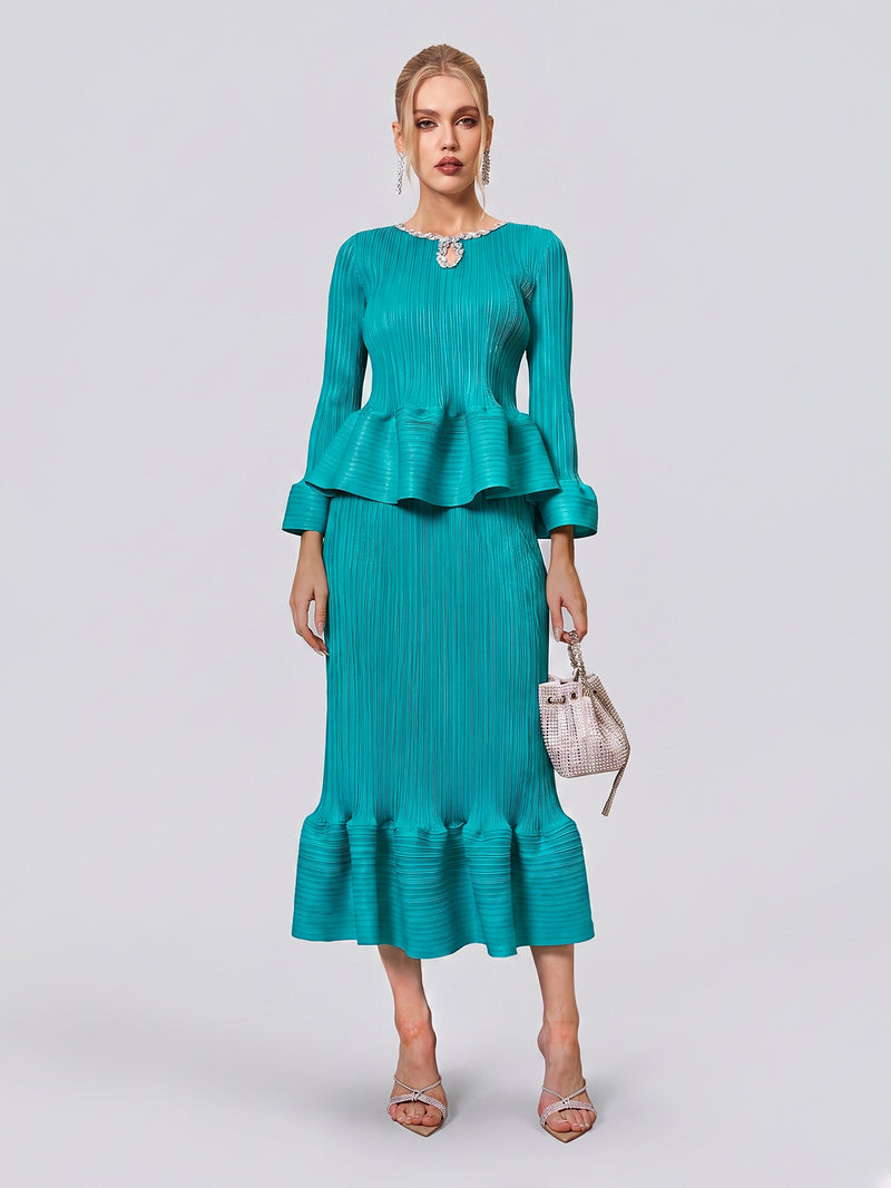 A woman stands against a plain background wearing the Social Queen Crystal-Embellished Pleated Peplum Set—a teal ensemble featuring a pleated peplum top and matching fishtail skirt. She accessorizes with a small handbag and silver sandals, making for a chic wedding guest look.