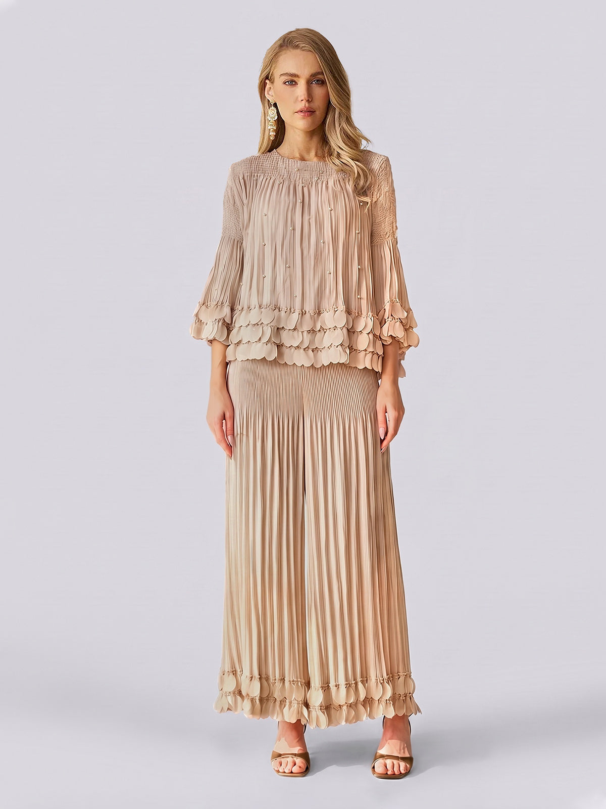 A woman stands against a plain light background, exuding effortless elegance in the Social Queen Layered Petal Beaded Pleated Set—a beige ensemble featuring a blouse with ruffled sleeves and beaded accents, paired with a matching long pleated skirt. This look is perfect for wedding guests.