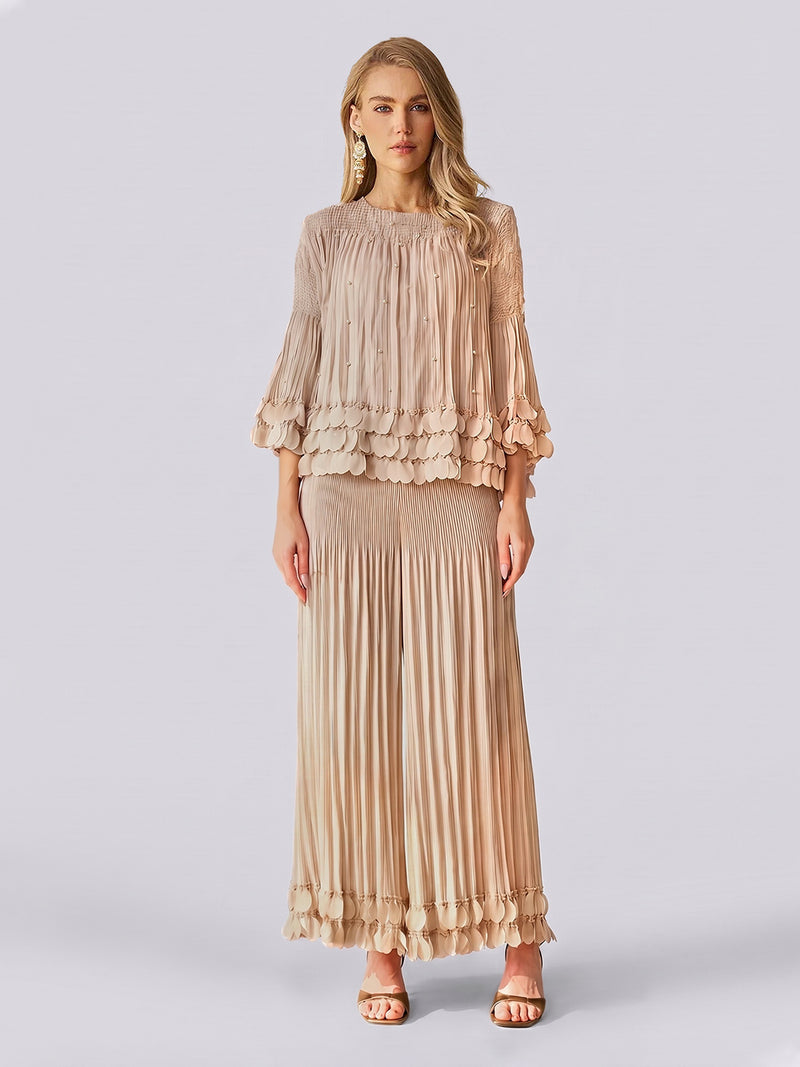 A woman stands against a plain light background, exuding effortless elegance in the Social Queen Layered Petal Beaded Pleated Set—a beige ensemble featuring a blouse with ruffled sleeves and beaded accents, paired with a matching long pleated skirt. This look is perfect for wedding guests.