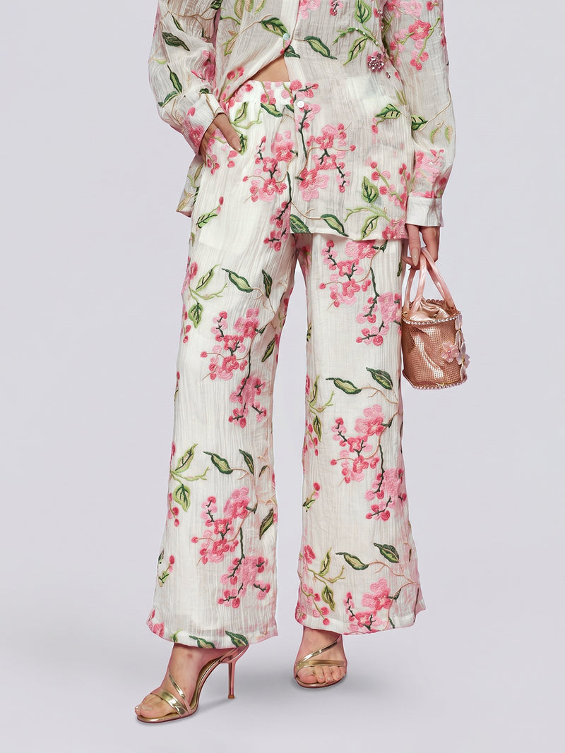 A person is wearing the Floral Embroidered Textured Wide-Leg Pants by Social Queen, paired with a matching long shirt featuring a pink floral pattern and delicate embroidery. They accessorize with a small shiny pink bag and gold high-heeled sandals.