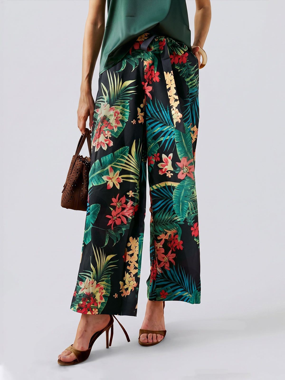 A person is standing against a plain background wearing Social Queen’s Tropical Print Wide-Leg Pants, paired with a green top, brown high heels, and a brown bag—the perfect ensemble for stylish vacation wear.
