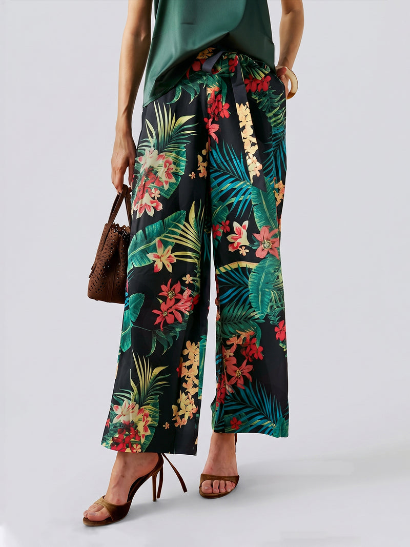 A person is standing against a plain background wearing Social Queen’s Tropical Print Wide-Leg Pants, paired with a green top, brown high heels, and a brown bag—the perfect ensemble for stylish vacation wear.