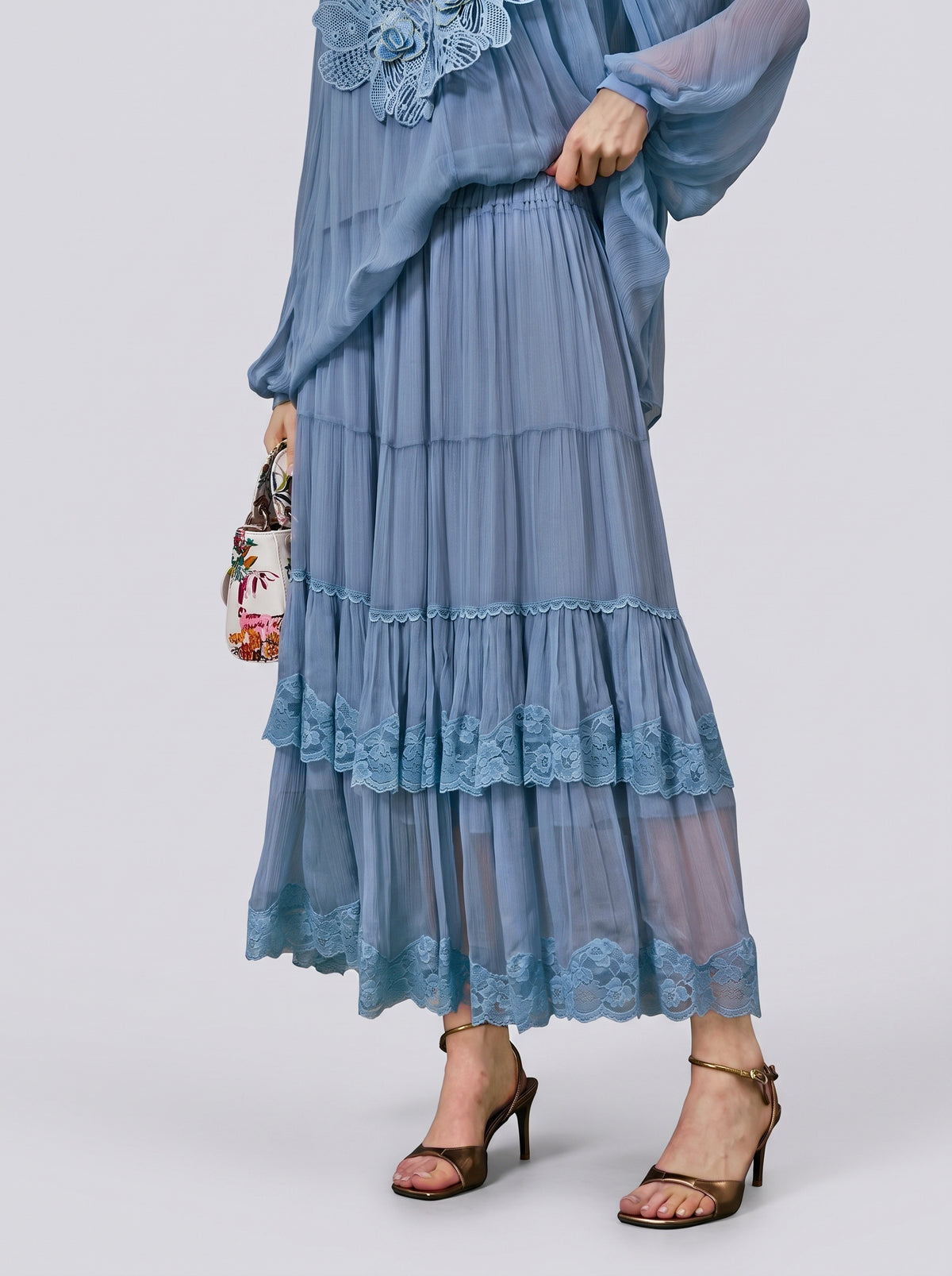 A person wearing the Social Queen Layered Lace Trim Skirt—a light blue, tiered chiffon design with floral lace trim and an A-line silhouette—paired with a matching top, brown high-heeled sandals, and carrying a small floral bag.