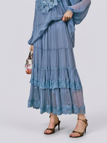 A person wearing the Social Queen Layered Lace Trim Skirt—a light blue, tiered chiffon design with floral lace trim and an A-line silhouette—paired with a matching top, brown high-heeled sandals, and carrying a small floral bag.