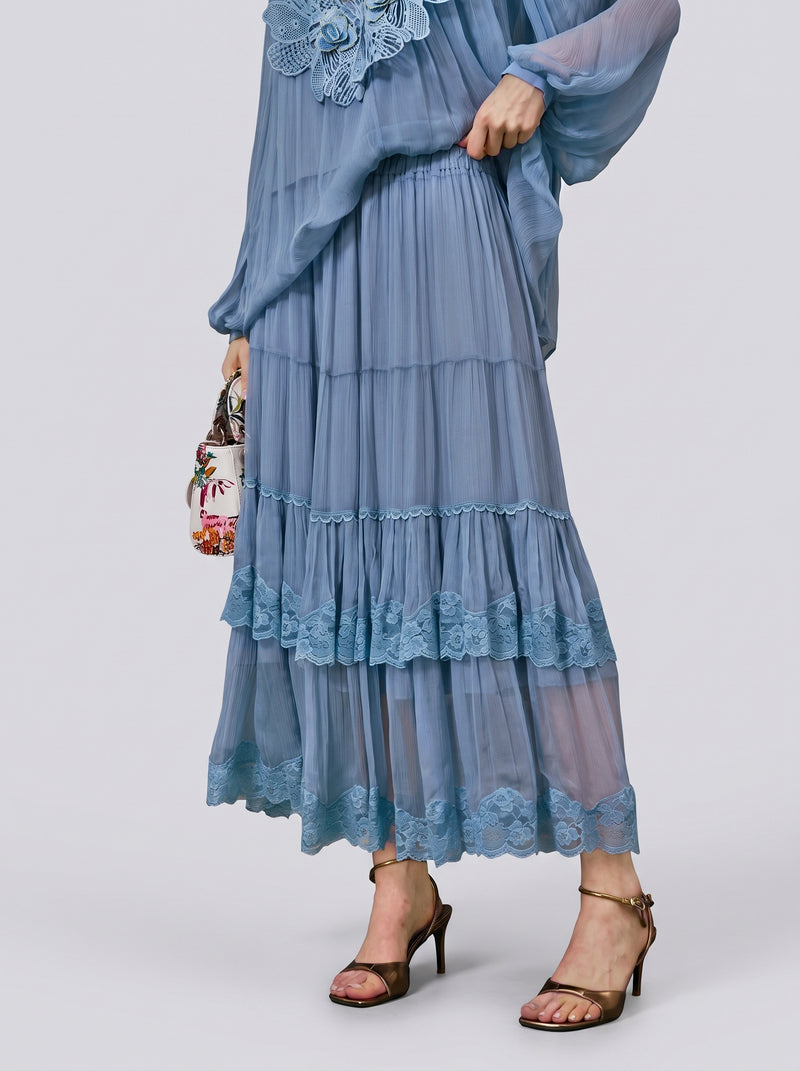 A person wearing the Social Queen Layered Lace Trim Skirt—a light blue, tiered chiffon design with floral lace trim and an A-line silhouette—paired with a matching top, brown high-heeled sandals, and carrying a small floral bag.