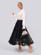 A woman in a white long-sleeve blouse and the Social Queen Jacquard Lace A-Line Midi Skirt, holding a small silver handbag and wearing strappy heels, stands against a plain light background. This elegant skirt features a high-waist A-line midi silhouette for timeless sophistication, making it an ideal choice for wedding guests.