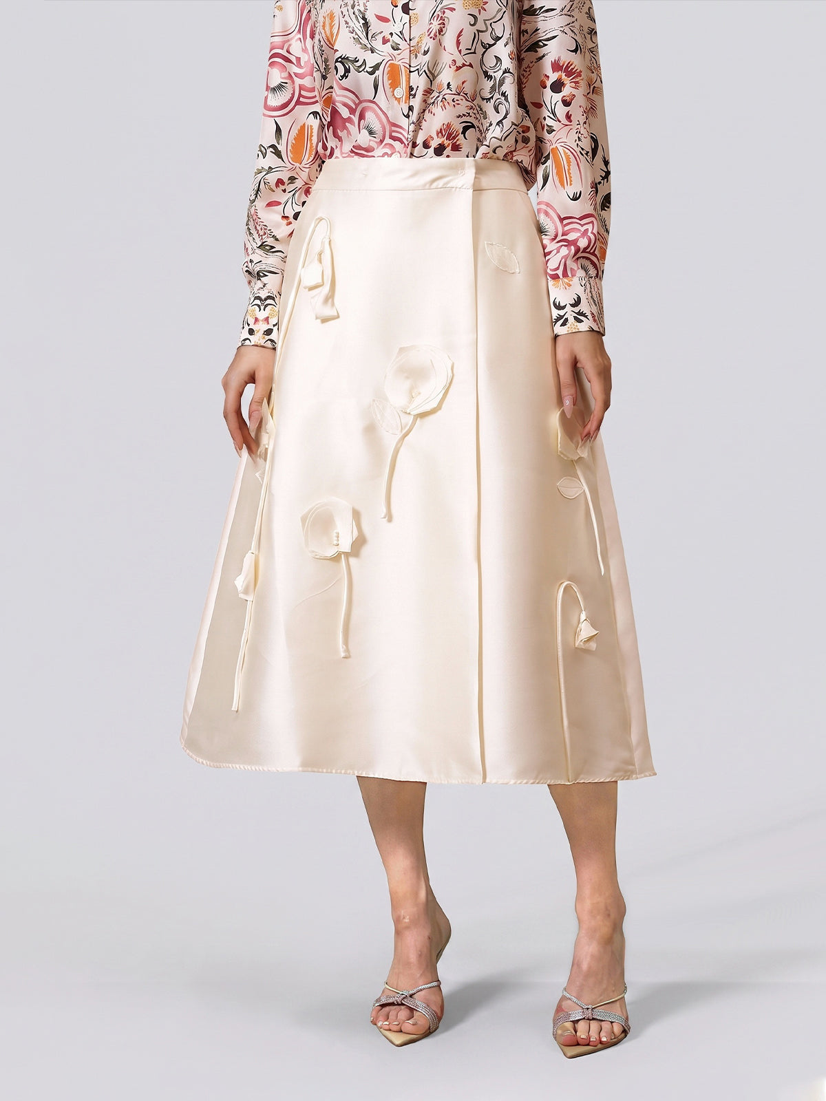 A woman stands against a plain background wearing the Social Queen 3D Floral Satin Midi Skirt—a long, cream midi skirt with intricate 3D floral appliqué—paired with a pink floral long-sleeve blouse and silver open-toe heels, perfect as an elegant choice for wedding guests or any sophisticated celebration.