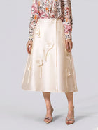 A woman stands against a plain background wearing the Social Queen 3D Floral Satin Midi Skirt—a long, cream midi skirt with intricate 3D floral appliqué—paired with a pink floral long-sleeve blouse and silver open-toe heels, perfect as an elegant choice for wedding guests or any sophisticated celebration.