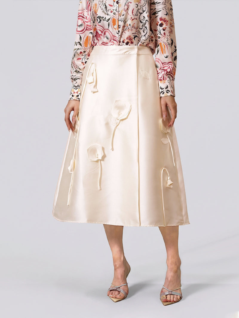 A woman stands against a plain background wearing the Social Queen 3D Floral Satin Midi Skirt—a long, cream midi skirt with intricate 3D floral appliqué—paired with a pink floral long-sleeve blouse and silver open-toe heels, perfect as an elegant choice for wedding guests or any sophisticated celebration.