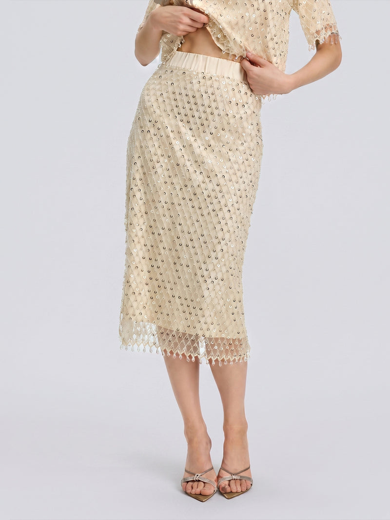 A woman stands against a plain background wearing the Social Queen Crystal Beaded Fringe Midi Skirt in beige, styled with a matching top. She poses with one hand lifting the shirt hem and the other resting on her hip.