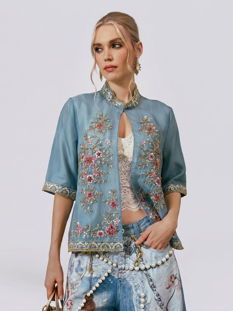 A woman wears the Social Queen Beaded Floral Embroidered Silk Jacket in light blue over a lace top and printed jeans, accessorized with a small handbag, against a plain background.